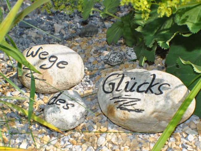 Steine mit der Aufschrift "Wege des Glücks" liegen auf Kies umgeben von Pflanzen