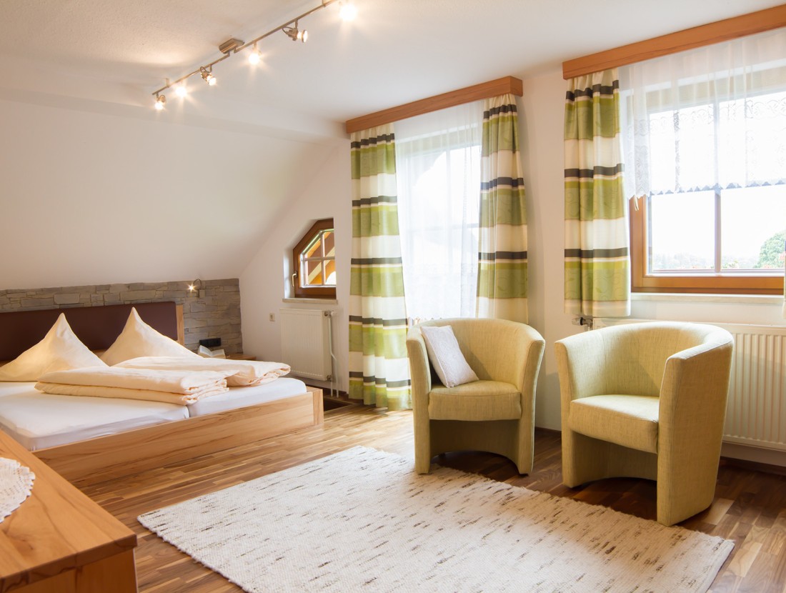 Helles Schlafzimmer mit Doppelbett, zwei grünen Sesseln und einem Fenster mit gestreiften Vorhängen